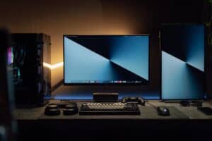 A dimly lit modern computer desk setup featuring dual monitors, a keyboard, and gaming accessories.