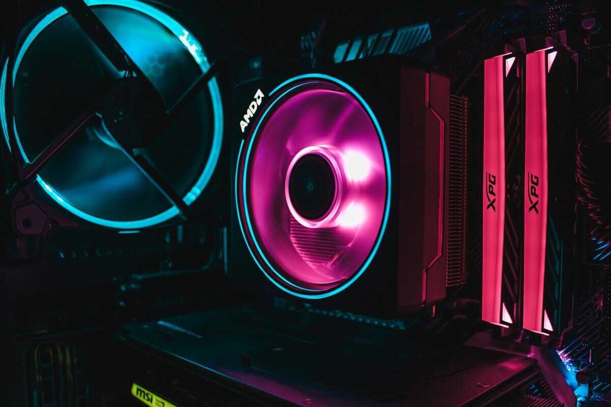 How to build a gaming PC close-up showing AMD RGB CPU cooler with pink lighting, XPG RAM modules, and cyan case fan on MSI motherboard
