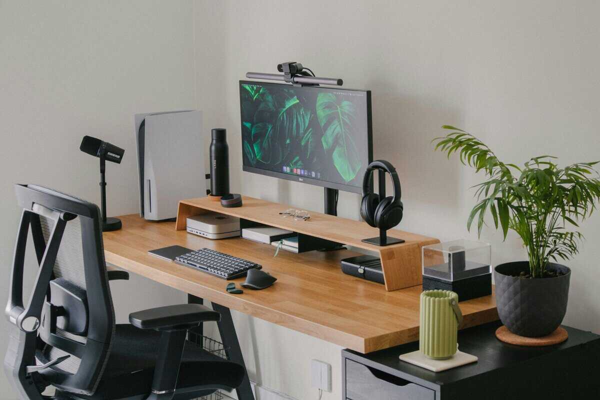 Organized gaming desk setup with wood surface and monitor riser, PS5 console, streaming microphone on boom arm, headphone stand, and ergonomic mesh chair
