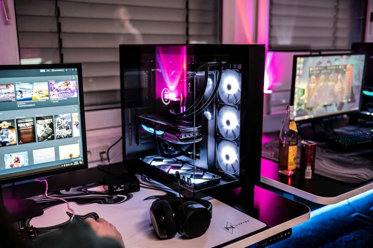 Best budget prebuilt gaming PC with tempered glass case, multiple RGB fans, dual monitor setup showing Steam library, and gaming headset on a deskmat
