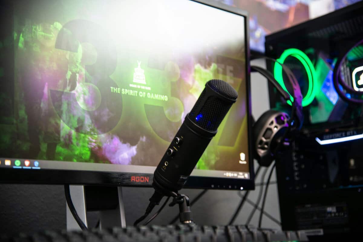 Best free recording software setup showing a USB condenser microphone on a tripod stand in front of an AOC AGON gaming monitor, with an RGB-lit gaming PC and headset in the background