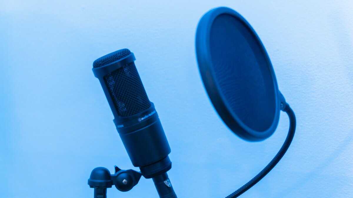 Black condenser microphone on a stand with a round nylon microphone pop filter attached via flexible gooseneck, showing proper placement for recording