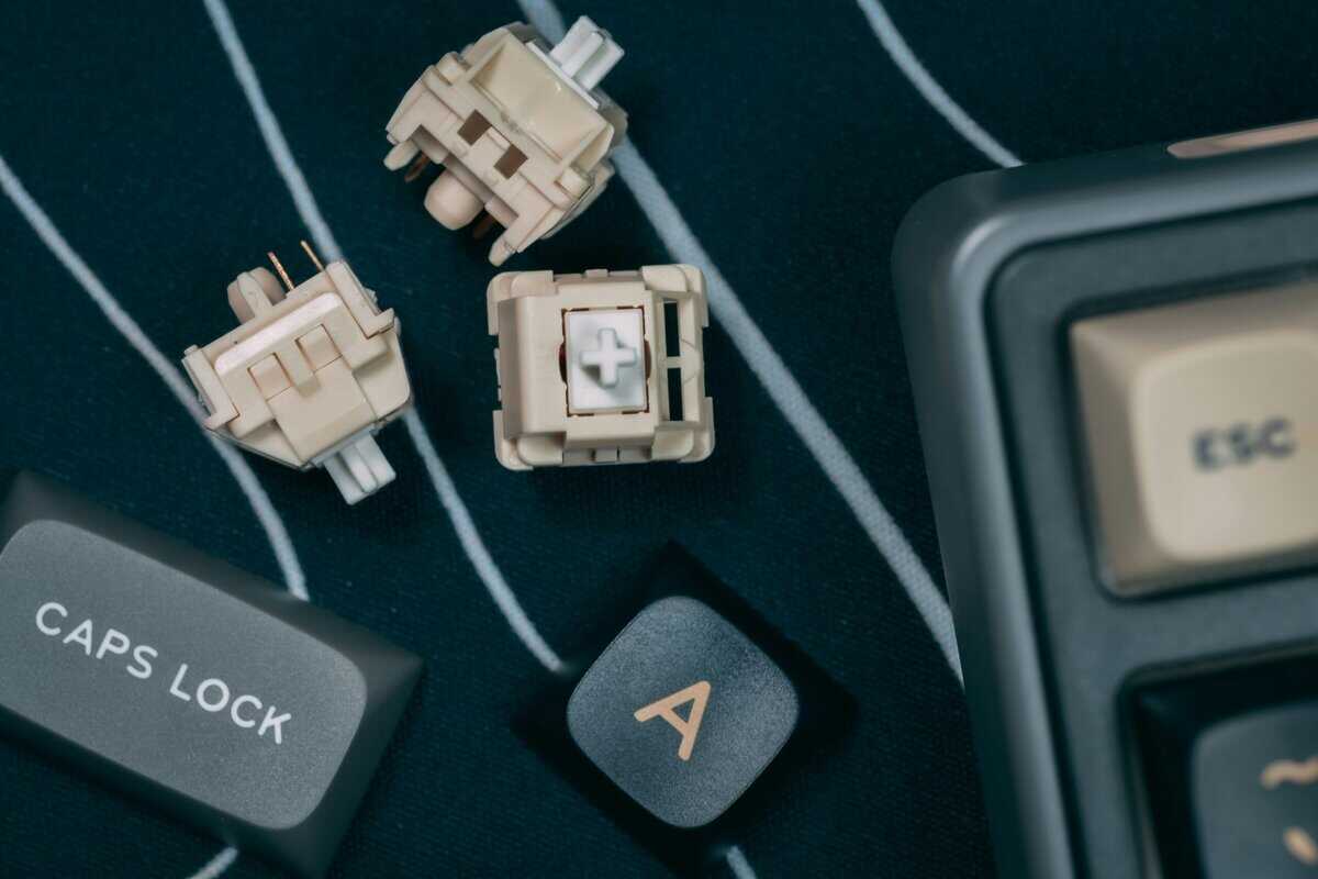 Three loose brown best tactile switches showing the stem and pin housing next to a mechanical keyboard on a dark deskmat