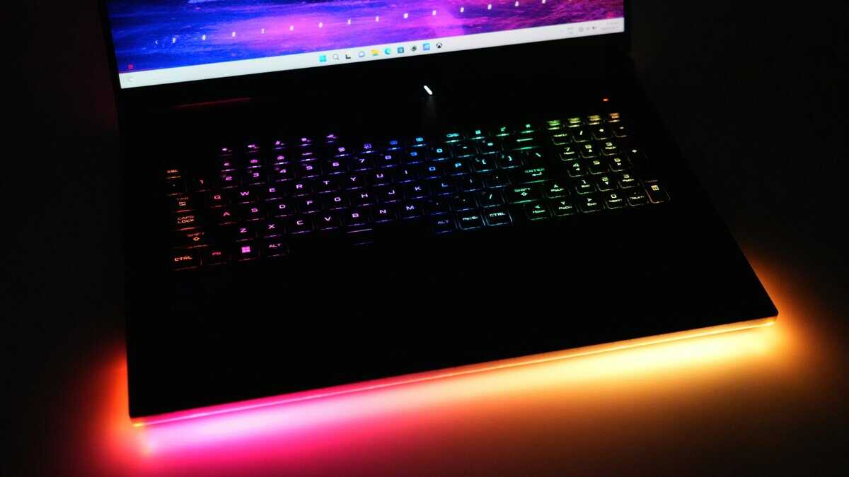 Gaming laptop open in a dark room with rainbow RGB keyboard backlighting and an orange LED light strip along the front edge, similar to a high-end RTX 5090 laptop