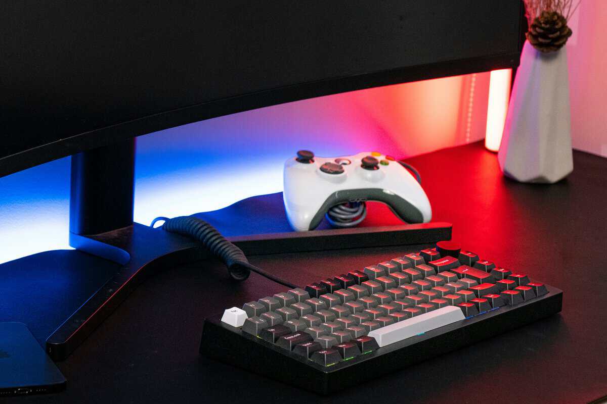 Compact mechanical keyboard with red and black keycaps next to an Xbox controller and an ambient-lit monitor, a typical setup that benefits from a regular how to clean a mechanical keyboard routine