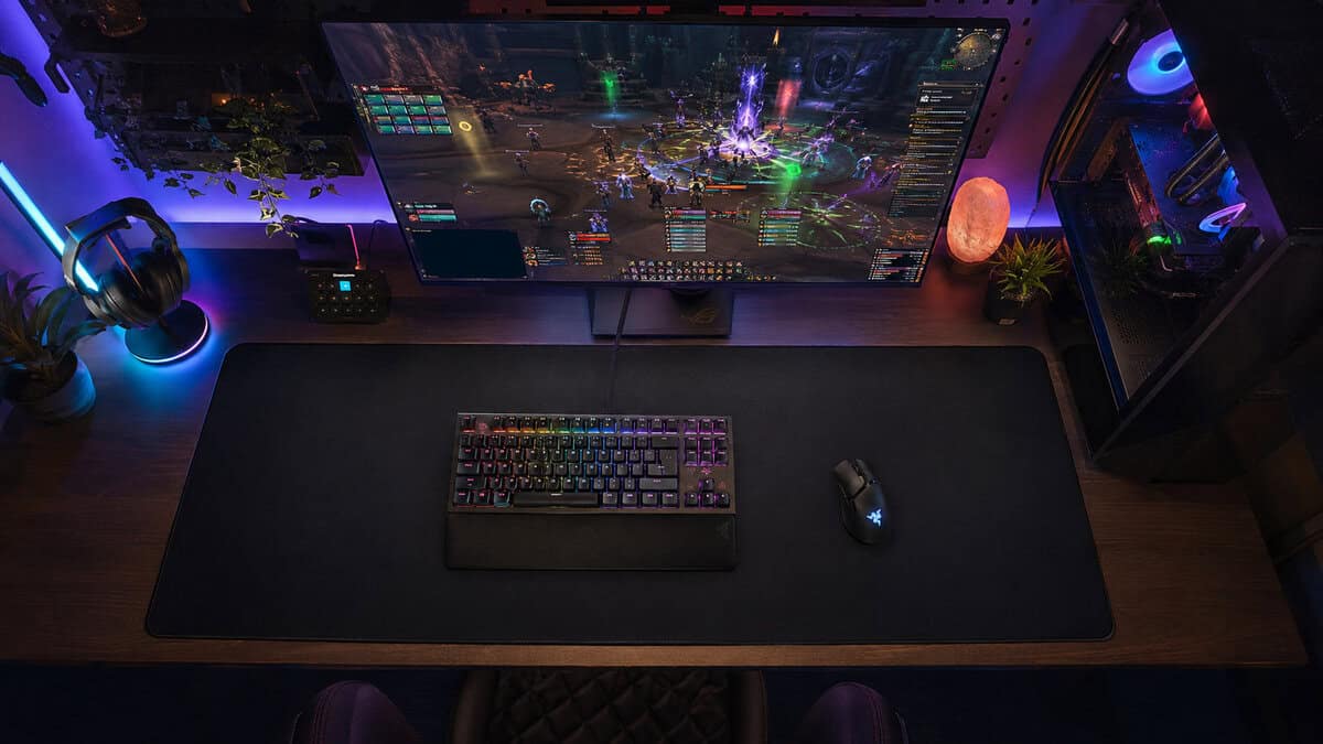 Top-down view of an RGB battlestation with a raid on screen and keyboard and mouse sharing one desk-scale pad, the setup the best MMO mousepad anchors
