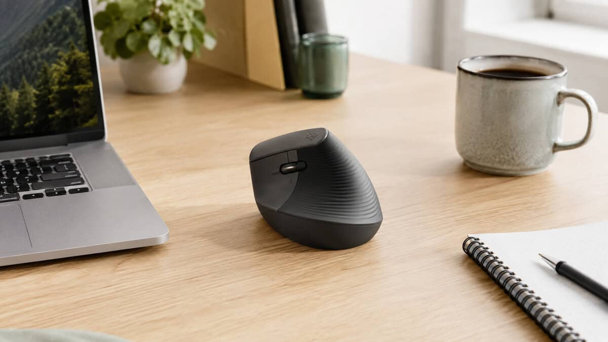 Compact black vertical mouse on a light wood desk next to a laptop, coffee mug and notepad, showing the smaller shell that suits the best vertical mouse for medium hands