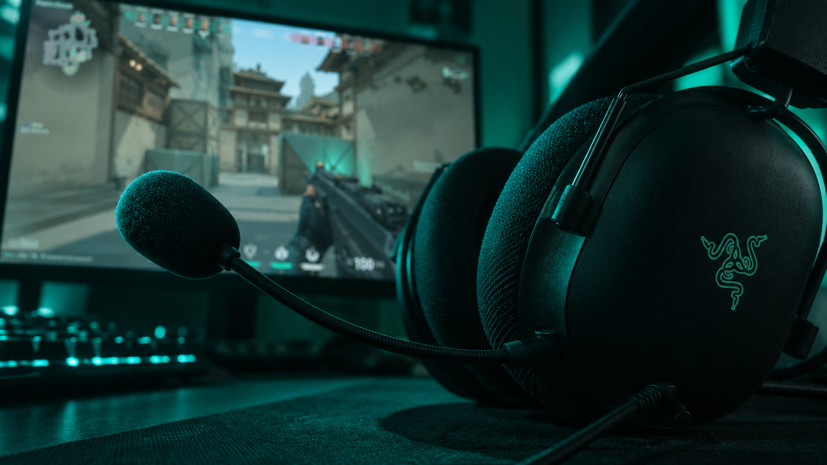 Close-up of a Razer BlackShark V2 X ear cup and boom mic in teal light with a Valorant map on the monitor behind it, the value pick for the best headset for Valorant