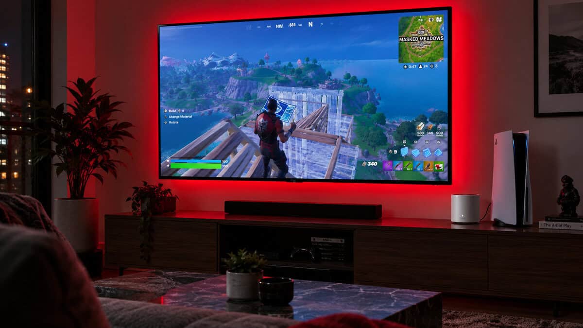 White TP-Link Deco X55 node on a media console beside a PS5 with a red-lit TV showing Fortnite, the living room end of the best mesh wifi system for a gaming home