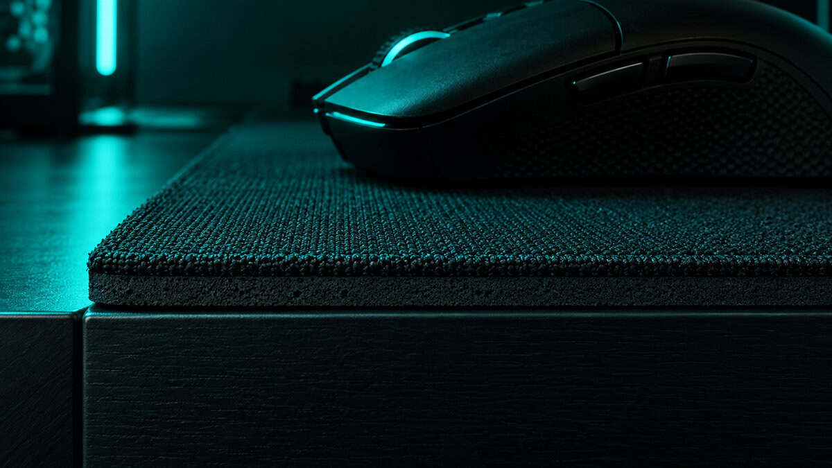 Side view of a black gaming mouse resting near the front edge of a thick cloth pad under teal light, showing the base thickness that makes the best mousepad for Tarkov comfortable over a long raid