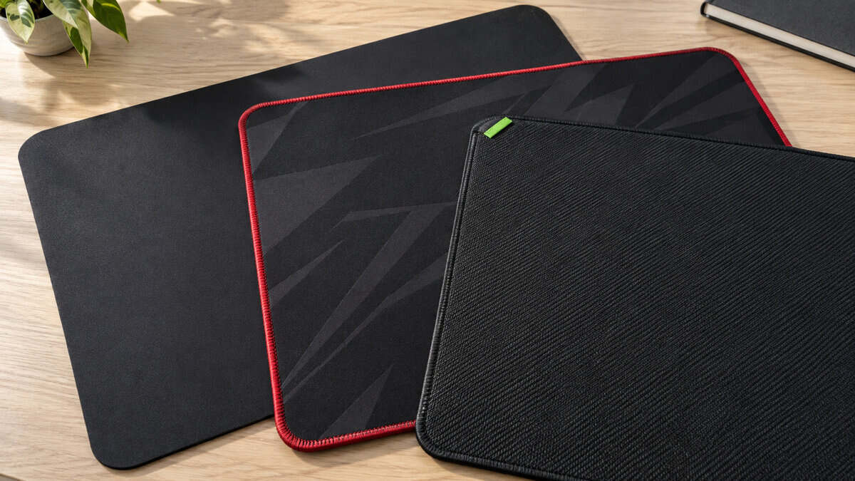 Three cloth gaming mousepads fanned on a light wood desk in daylight, a plain black pad, a red-stitched ROG-style pad, and a textured Razer-style pad, the three picks for the best mousepad for Wardogs