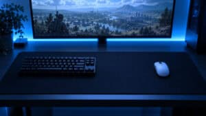 Wide extended black cloth mousepad under a tenkeyless keyboard and a white wireless mouse, with an open-field shooter landscape on the monitor and cool blue lighting, the setup the best mousepad for Wardogs is built around