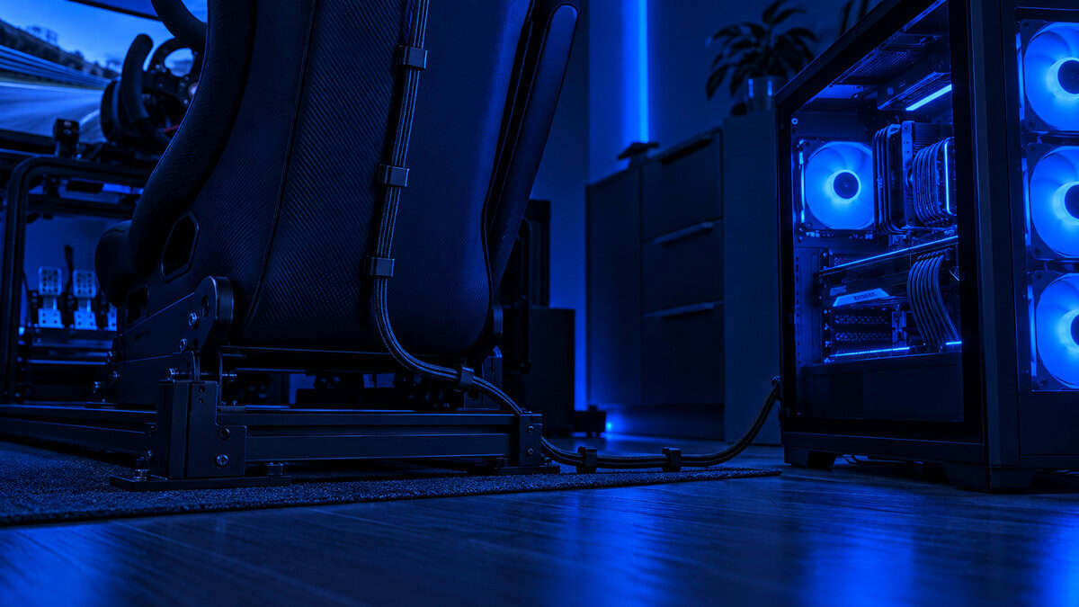 Low-angle view along the floor of a sim rig under deep blue light, a USB-C cable routed up the back of a bucket seat and down to a mid-tower PC with glowing fans, the wired tether the best VR headset for sim racing depends on