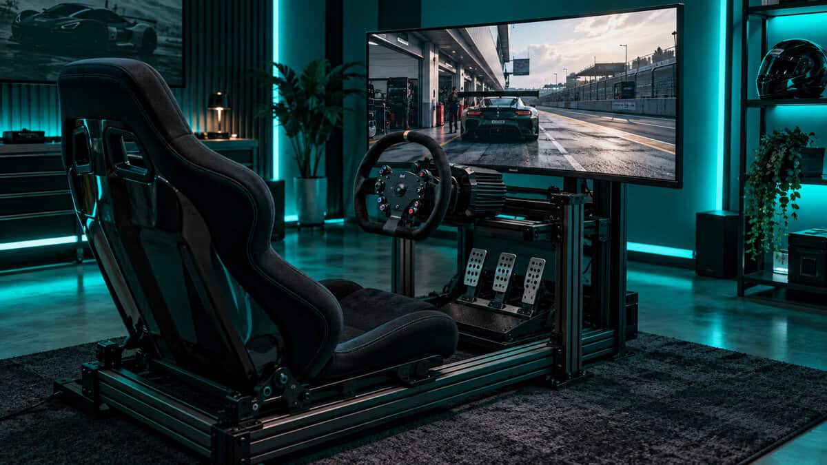 Fixed aluminum sim racing cockpit with a reclining bucket seat on sliders and a direct-drive wheel, facing a monitor showing a car in the pit lane under teal room lighting