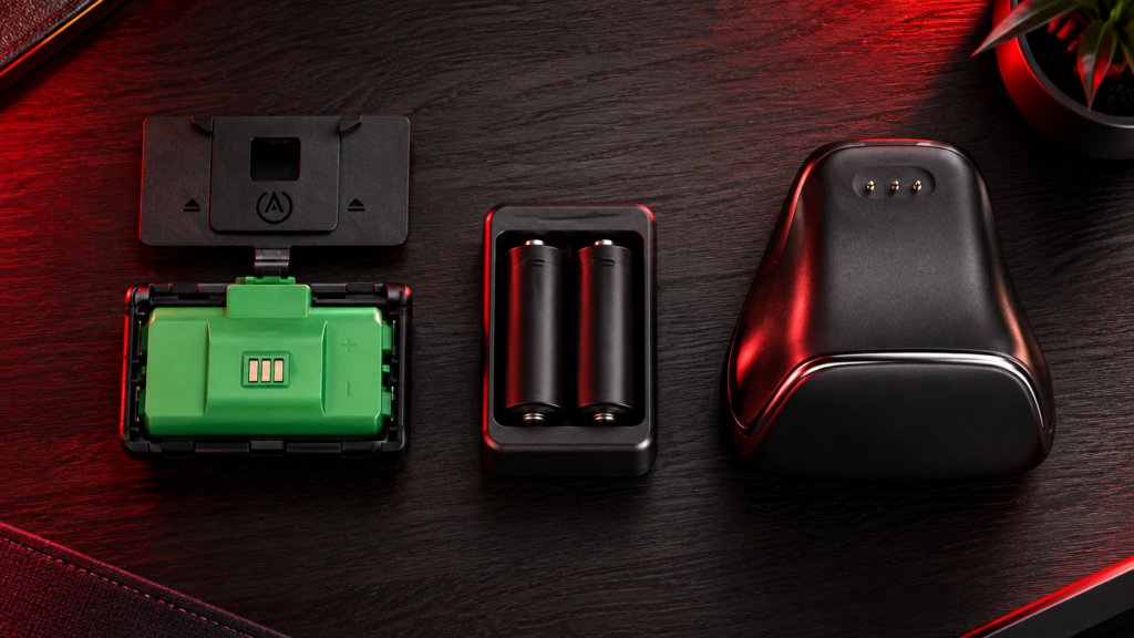 Top-down comparison of a green rechargeable controller battery pack with its door, two AA batteries in a wall charger and a charging stand on a dark desk in red light, three kinds of Xbox controller charger