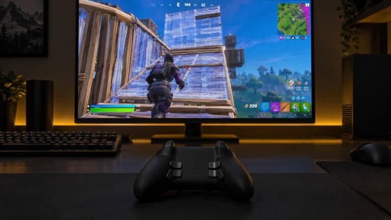 Black controller with four rear paddles resting on a dark desk in front of a monitor showing a Fortnite build fight, a golden yellow light strip glowing behind the screen, set up as the best controller for Fortnite