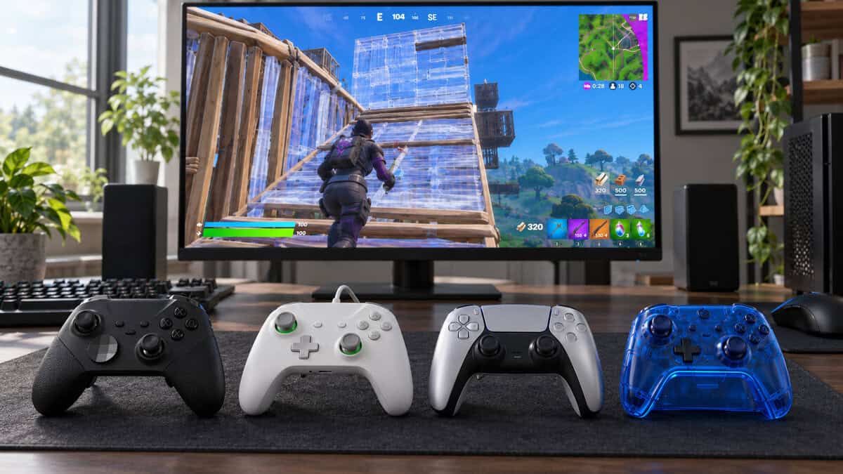 Four controllers in black, white, silver and clear blue lined up on a desk mat in window daylight, comparing options for the best controller for Fortnite, with a Fortnite build fight on the monitor