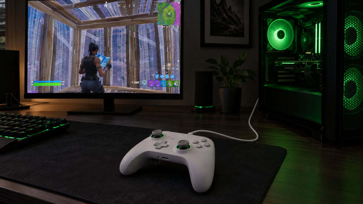 White wired controller on a black desk mat with its cable running to a PC tower lit by green fans, a budget route to the best controller for Fortnite, with a box-up build on the monitor