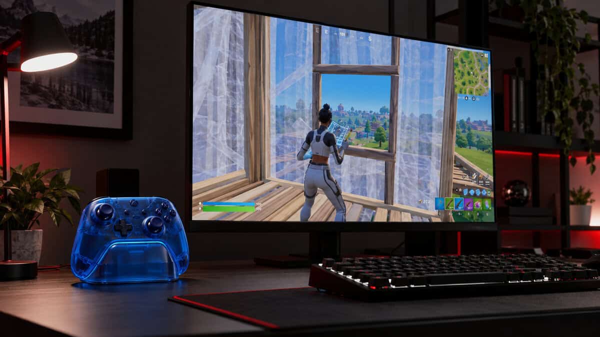 Transparent blue wireless controller standing in its charging dock beside a desk lamp and red shelf accents, a cable-free take on the best controller for Fortnite, with a wall edit in Fortnite on the monitor