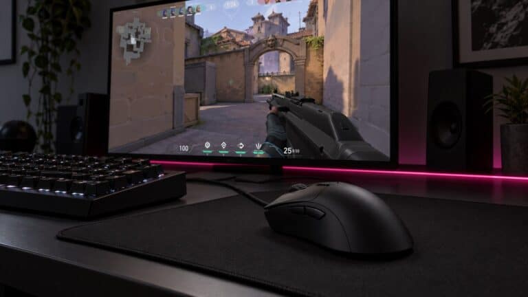 Black right-handed ergonomic wired mouse with a long sloped back on a black cloth pad beside a keyboard, an example of the best mouse for claw grip, with Valorant on the monitor