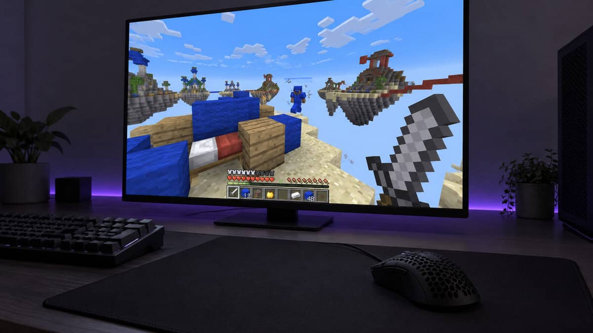 Black lightweight honeycomb gaming mouse on a large cloth pad in front of a monitor showing a Minecraft Bedwars fight on sky islands, a violet light strip glowing behind the screen, a PvP desk built around the best mouse for Minecraft
