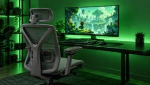 A gray mesh herman miller alternative with a headrest and padded armrests faces a green-lit gaming desk where an ultrawide monitor shows a cartoon forest adventure