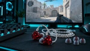 A pair of red translucent iems for gaming with a braided silver cable and spare ear tips on a desk mat, Counter-Strike 2 on the monitor behind them