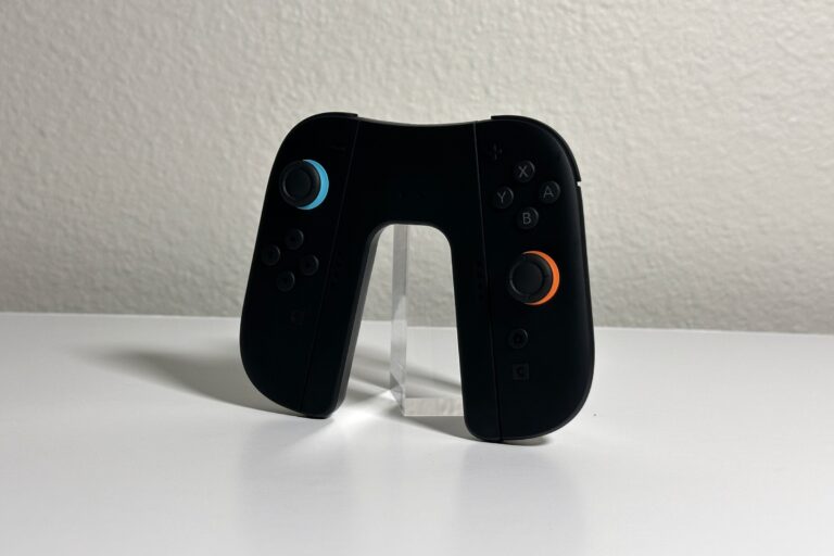JSAUX Switch 2 charging grip holding a black pair of Joy-Con 2 with blue and orange stick rings, angled on a white desk