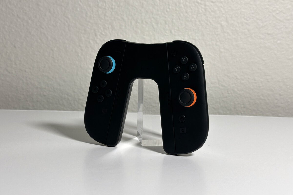 JSAUX Switch 2 charging grip holding a black pair of Joy-Con 2 with blue and orange stick rings, angled on a white desk