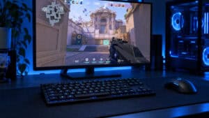 Black tenkeyless low-profile mechanical keyboard with blue backlighting on a dark desk mat, with a monitor showing Valorant gameplay behind it